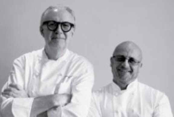 Two chefs wearing chef's whites, with arms folded.