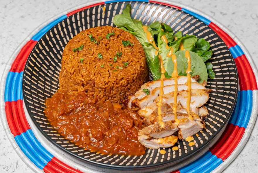 Dish of African food including sliced meat, rice and stew on a plate with black geometric pattern.