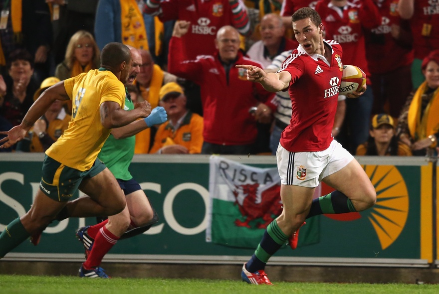Rugby players from the Wallabies in yellow and green and from the Lions in red and white, playing on a green field, with fans in the stadium cheering.