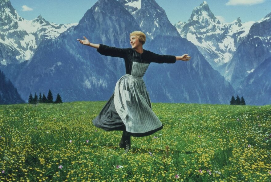 Classic still from The Sound of Music, Julie Andrews standing in a green field surrounded by snowcapped Alps, her arms oustretched.