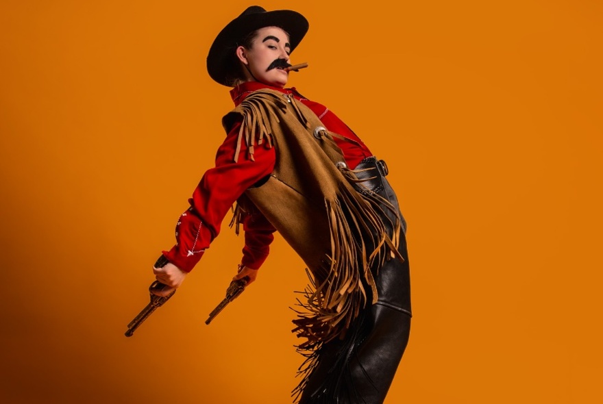 Comedian, Lil Wenker, dressed as a Wild West cowboy, posing with chest and hips pushed forward and pistols held out behind her.