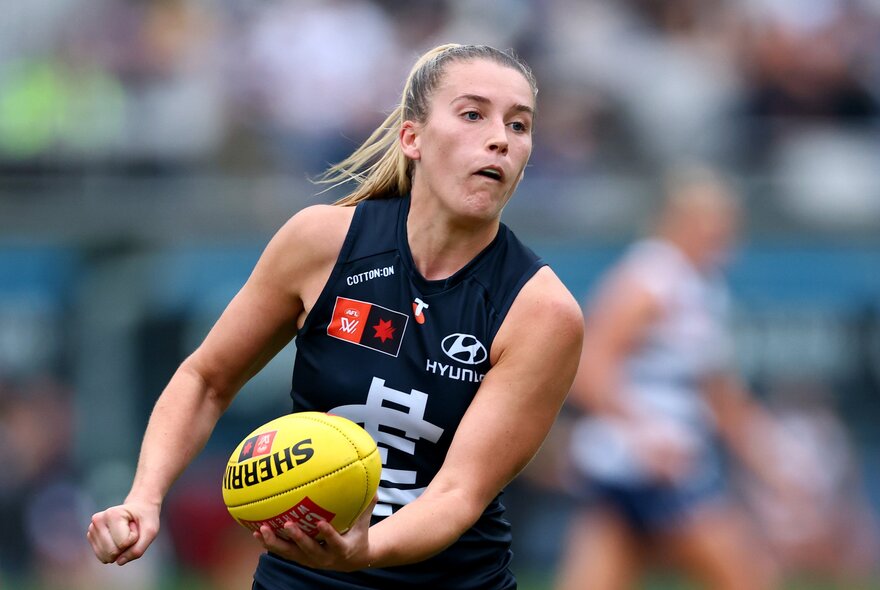 An AFLW player in a Carlton jersey handballing a yellow football during a game.