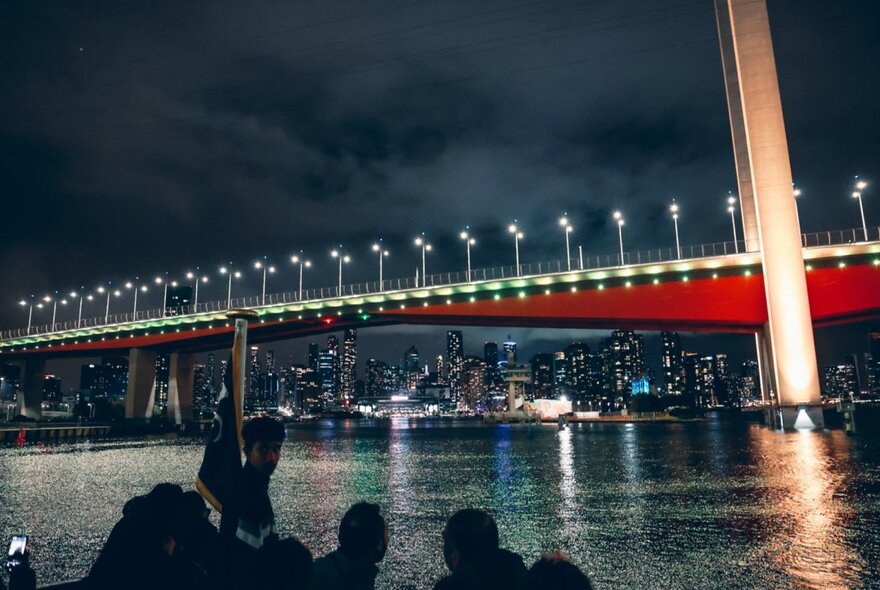 People viewing the Bolte Bridge in Melbourne, lit up at night, from a boat on the Yarra River.