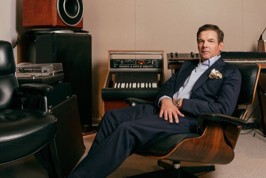 Musician wearing a suit leaning back in an Eames chair surrounded by vintage audio equipment.