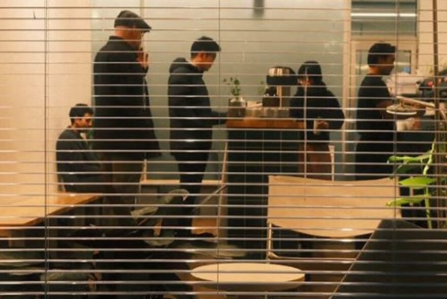 Looking into the interior of a small cafe from the outside, the window has thin venetian blinds and people are waiting in line ot order a coffee.