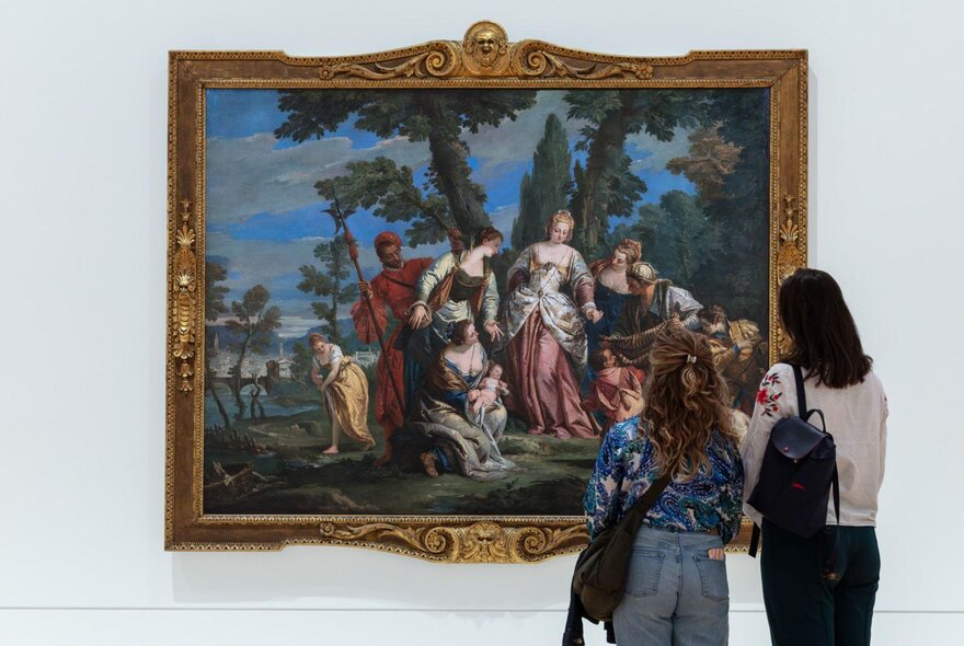 The back of two people standing and looking at an 18th century oil painting in a heavy gold frame hanging on a white gallery wall.