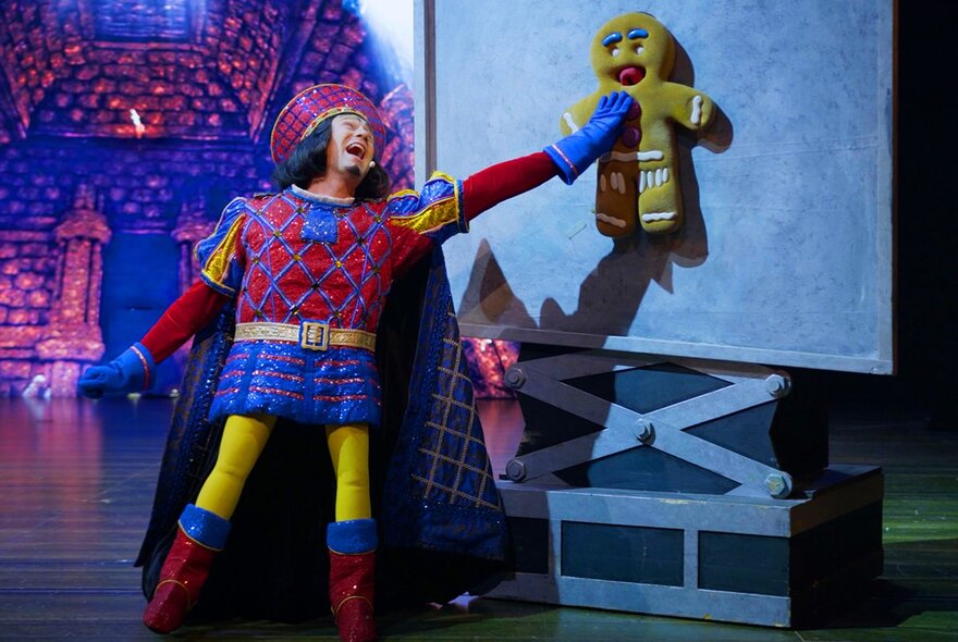 The Prince character in Shrek The Musical holding up the gingerbread man cookie and laughing, on a theatre stage.