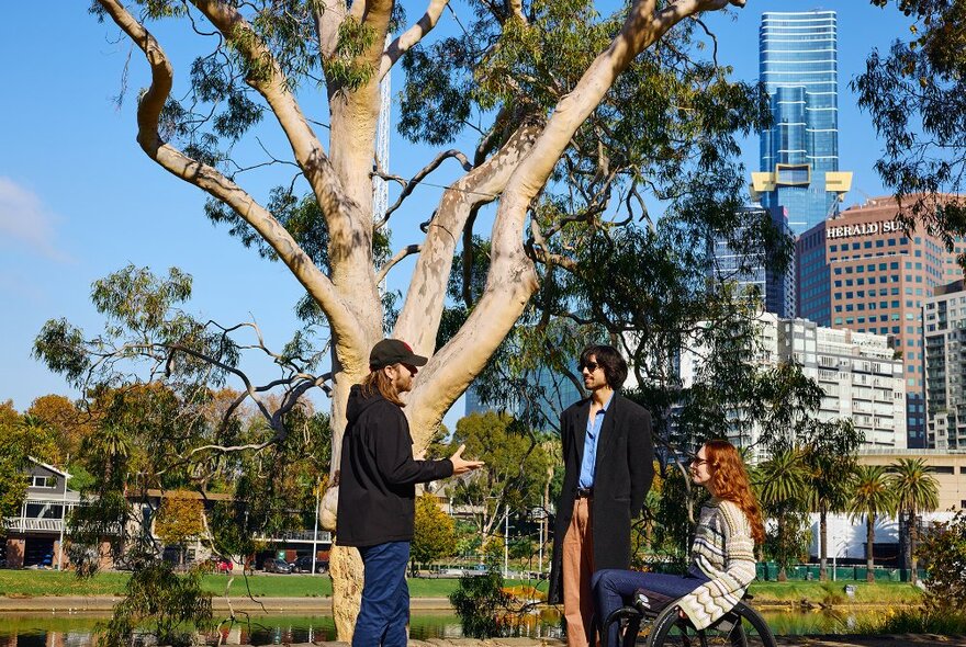 A guide talking to two people beside a tree and river, the woman is using a wheelchair.