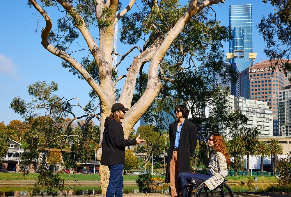 A guide talking to two people beside a tree and river, the woman is using a wheelchair.
