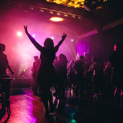 People dancing at a nightclub with pink glowing light, a woman with her arms in the air. 