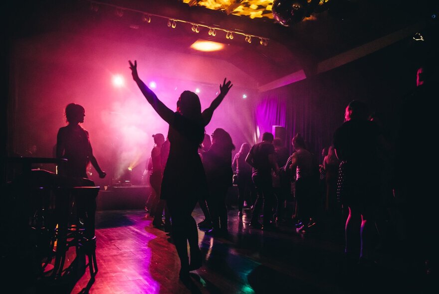 People dancing at a nightclub with pink glowing light, a woman with her arms in the air. 