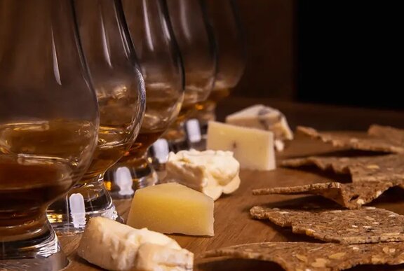 A sample of five whiskey pours with a small chunk of cheese and a cracker in front of each glass.