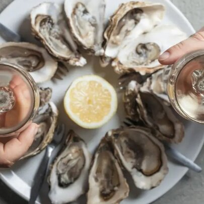 Two people's hands holding white wine glasses over a plate of natural oysters, seen from above.