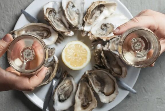 Two people's hands holding white wine glasses over a plate of natural oysters, seen from above.