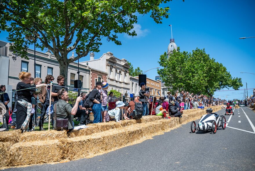A billy cart race with onlookers standing behind a hay bail wall