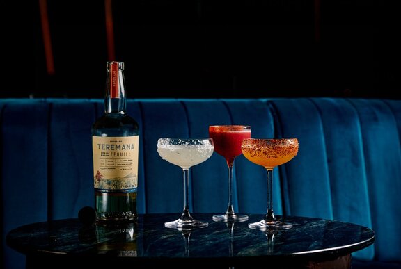 A tequila bottle next to three different kinds of margaritas on a marble table in front of blue velvet couches.