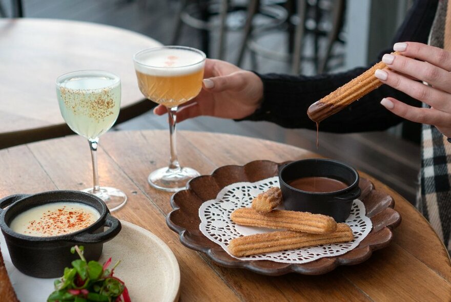 Someone dunking a churro into melted chocolate while holding a cocktail.