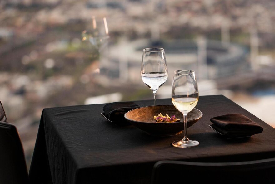 A small table covered in a black cloth, with two wine glasses and a plate, positioned against floor to celling windows.
