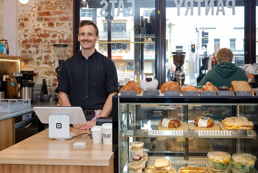 A man standing behind a Square terminal read to serve customers in a cafe with a display case featuring rolls. 