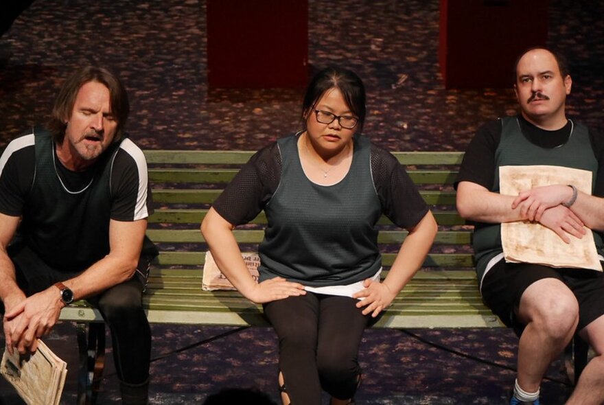 Three actors on stage performing an improvised theatre routine, all of them seated on a bench seat.