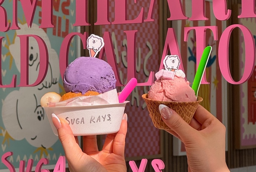 Two small bowls of ice cream being held up in front of window with pink signage.