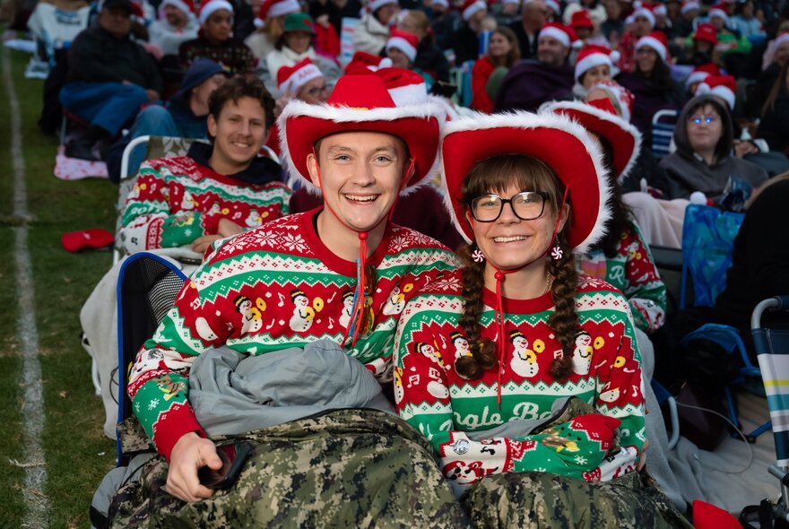 A couple smiling at an outdoor event while wearing matching festive red and green Christmas jumpers and red cowboy hats.