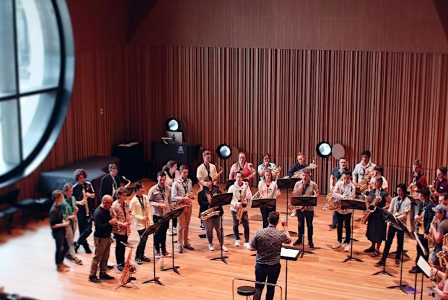 A standing ensemble of saxophone players with a conductor leading them, performing in a concert hall.