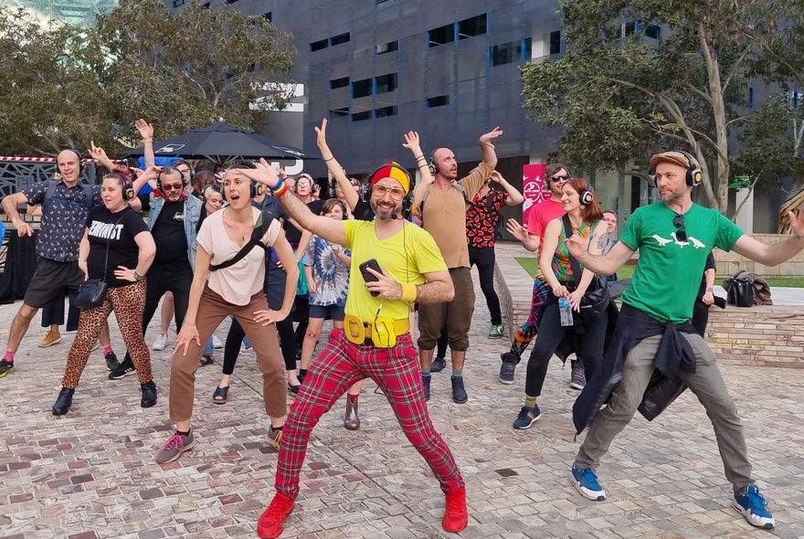 A group of people taking part in a silent disco dance-walk, wearing headphones with a host tour guide leading the dance moves, in Federation Square's main plaza.