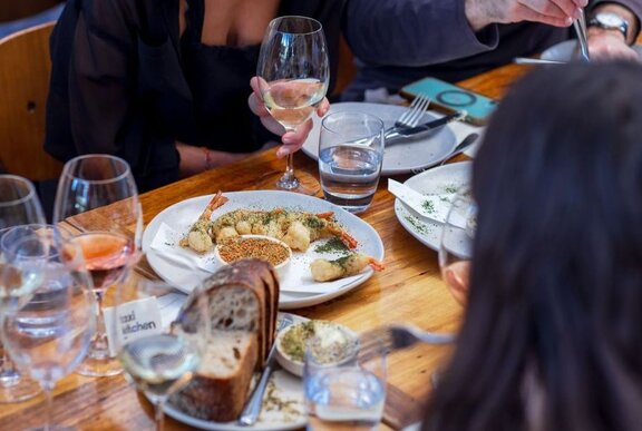 A table in a restaurant with plates of food mid-meal, bread and glasses of wine.