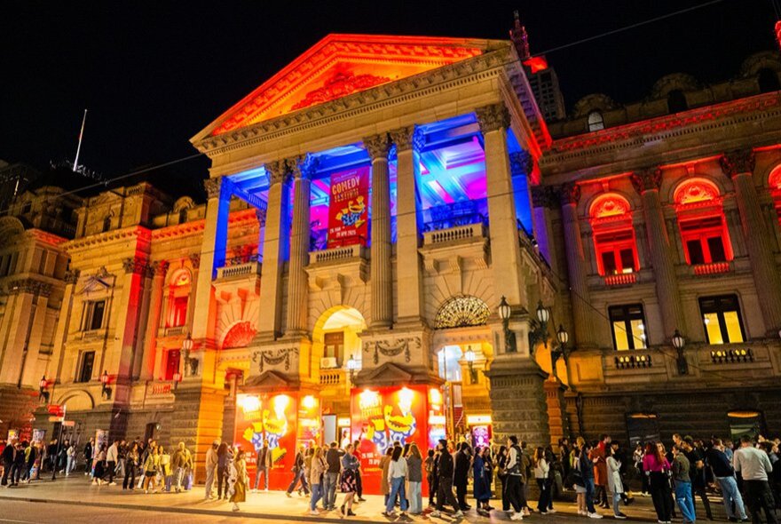 The Town Hall at night with Melbourne International Comedy Festival banners, signage, and red and blue spotlights and people lined up in queues on the footpath.