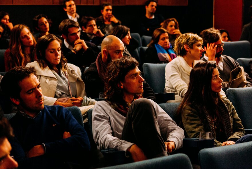People seated in a lecture room, listening to something out of sight on the stage in front of them.