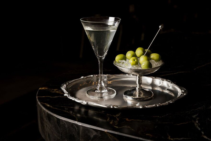 A classic martini served on a silver tray alongside a small bowl of green olives on ice, against a black background.