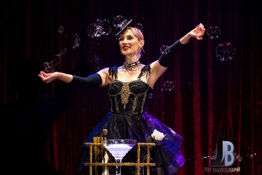 A cabaret artiste waving her arms in front of a large cocktail glass, wearing black corset and hat.