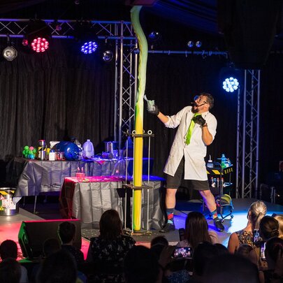 A man in a white lab coat on stage doing experiments in front of an audience. 