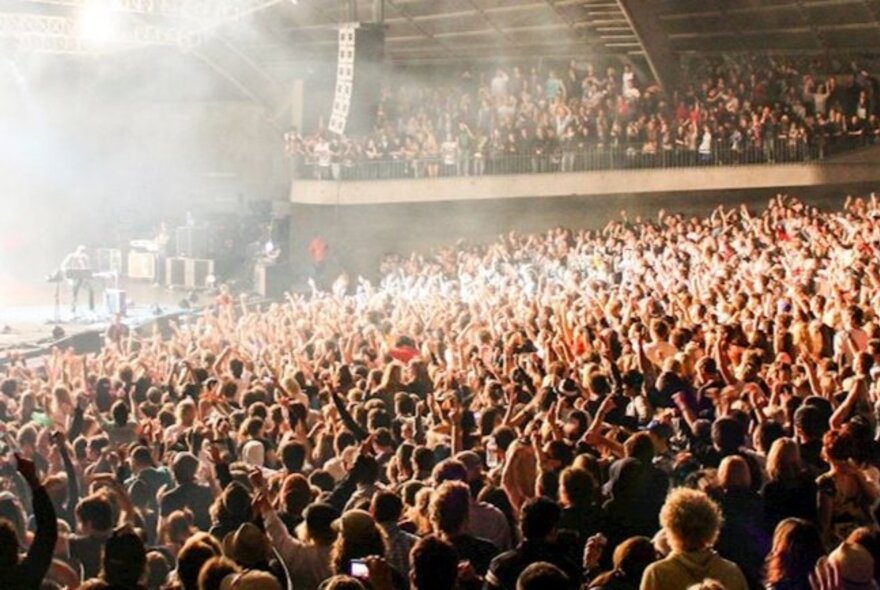 A massive crowd at the Sidney Myer Music Bowl, all standing with their hands in the air as a musician on stage performs.