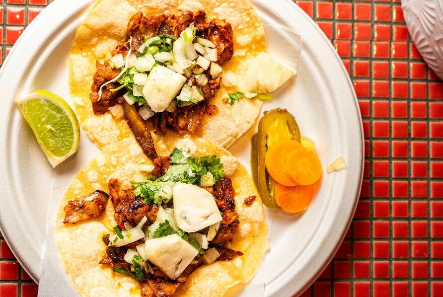 Overhead view of a plate that has two open tacos on it, pickles and a lime garnish, on a tiled red table top.