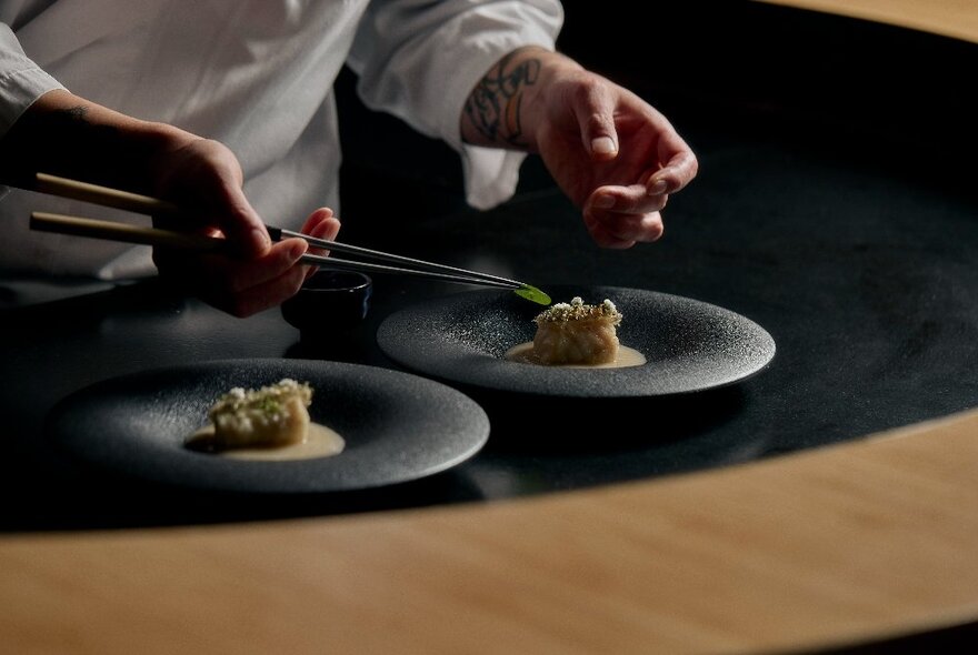 Chef's hands holding chopsticks, arranging food delicately on black plates.