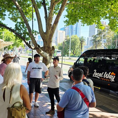 A group of people in a circle listening to a speaker under a tree with a black van parked next to them.