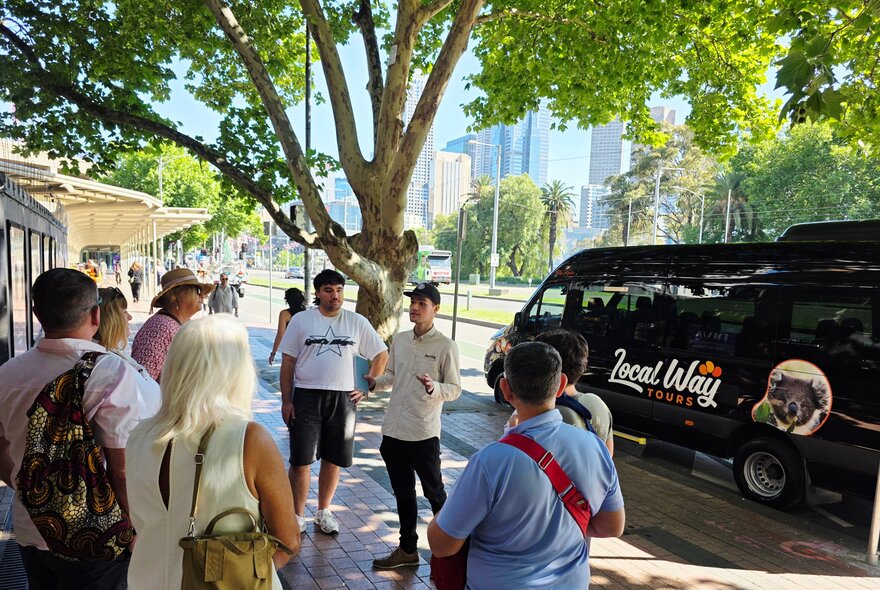 A group of people in a circle listening to a speaker under a tree with a black van parked next to them.