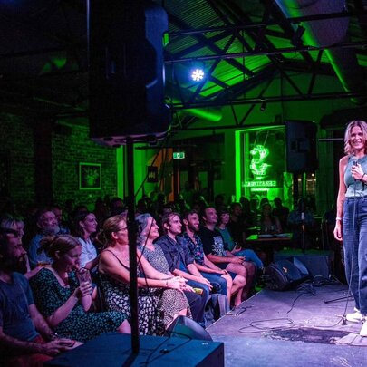 A woman on stage in front of an audience on a small venue with green lighting in the background. 
