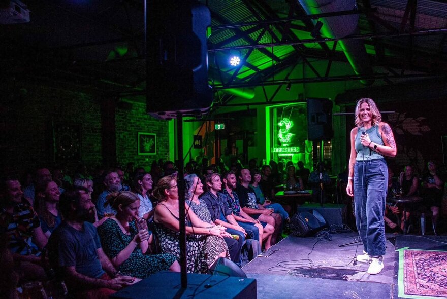 A woman on stage in front of an audience on a small venue with green lighting in the background. 