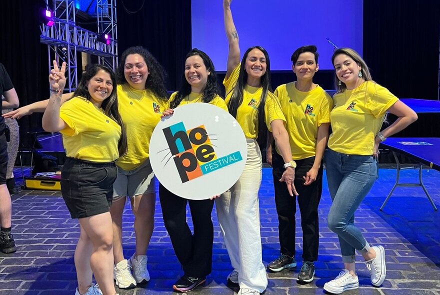 Six volunteers at the Hope Festival wearing matching yellow t-shirts and holding up a round sign with the words HOPE Festival written on it.