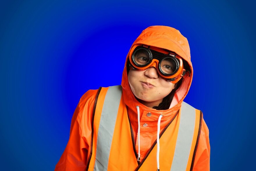 A person wearing a high vis orange raincoat with the hood raised over their head, orange goggles covering their eyes, and expanding their cheeks with air, standing against a blue background.