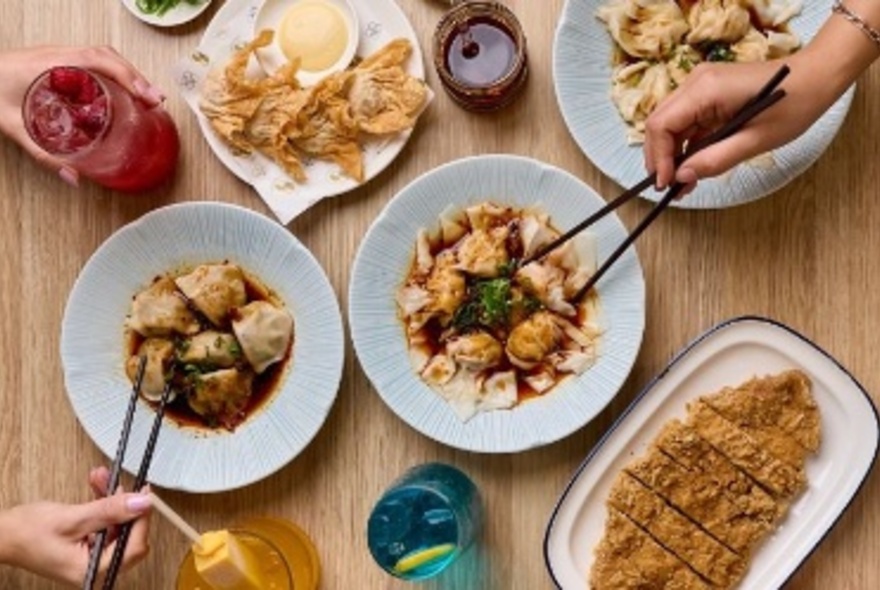 Hands holding chopsticks skewering dumplings from plates of Chinese food on a table with drinks and other dishes.