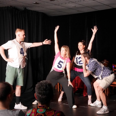 An ensemble cast of impro actors on a stage, performing in front of a live audience in a small theatre.