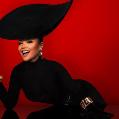 A woman with an outrageously large black hat dressed in a black outfit smiling and holding a glass against a deep red background. 