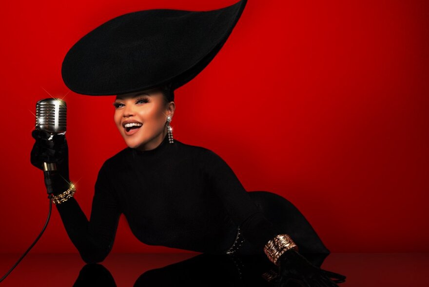 A woman with an outrageously large black hat dressed in a black outfit smiling and holding a glass against a deep red background. 