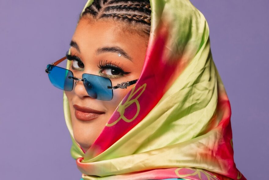 Performer Milo Hartill with blue sunglasses, her face angled towards the viewer, wearing a headscarf around her braided hair.