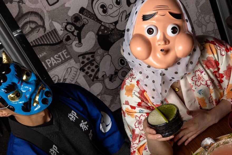 Two people wearing Japanese theatrical face masks, posed in Japanese robes inside a restaurant.