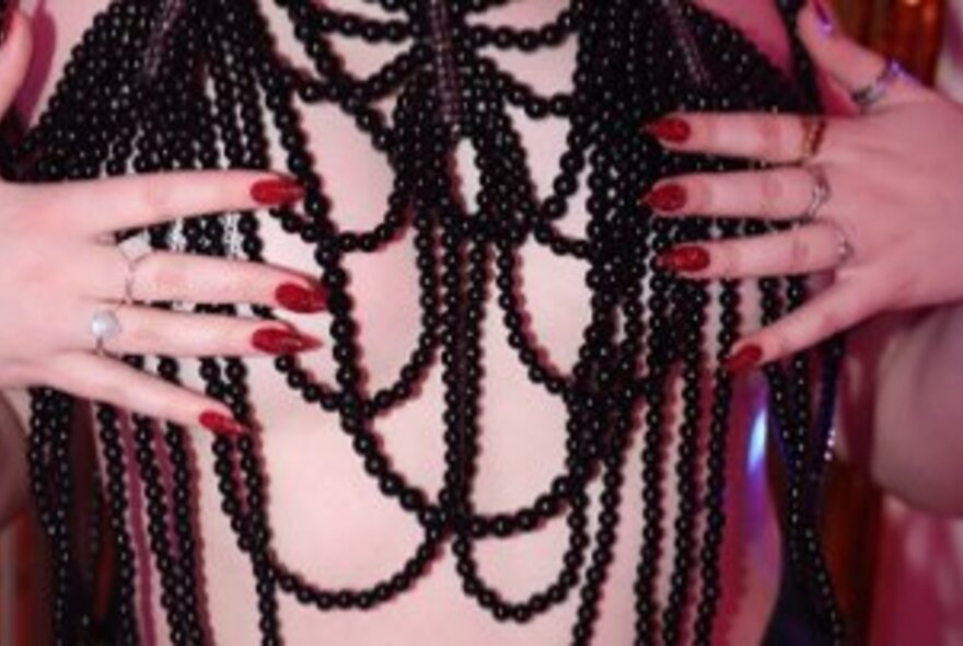 Hands wearing red nail polish positioned in front of a naked torso draped in black beads.